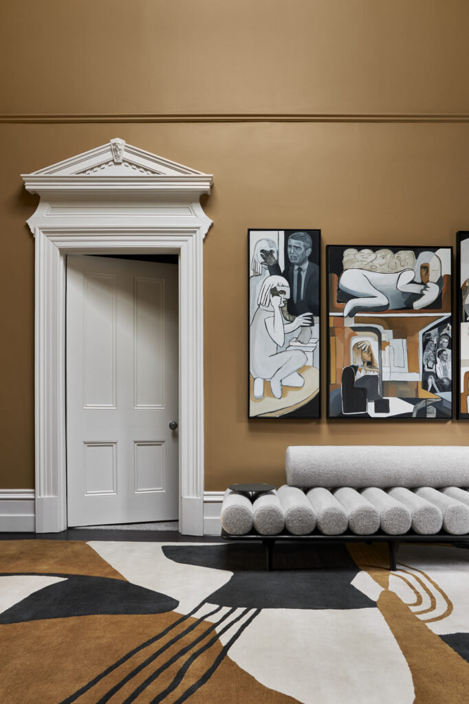 Simone Haag The Whiskey Room hall with mustard walls with art hanging on the walls and with a cream white door and with a grey chaise long and with a rug