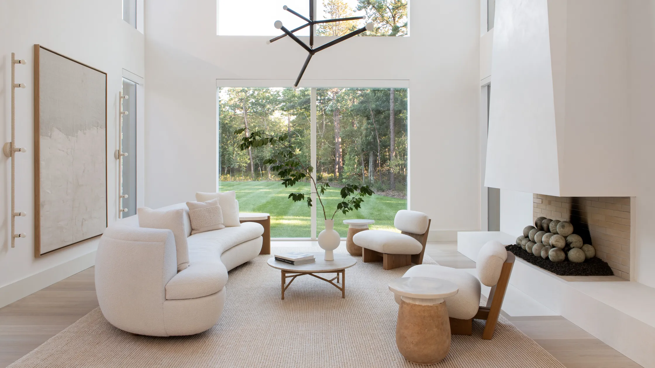 Minimalist Interior Design living room with large windows and with white walls and with a curved white sofa and with white boucle armchairs and with a stone center table and with wooden side tables and with a jute rug