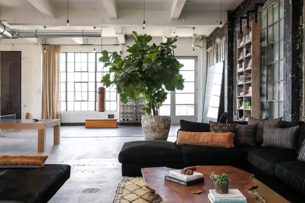 Raw Aesthetics in a open space with concrete floors and with brick walls and with beams on the ceiling and with a leather sectional sofa and with a wood center table and with a large travertino vase