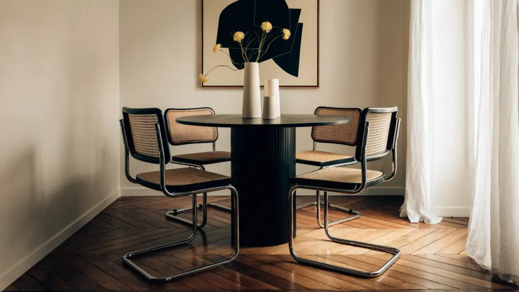 small apartment decor dining area with cream walls and with wood floors and with a black round dining table and with mid-century modern dining chairs and with a painting framed on the wall