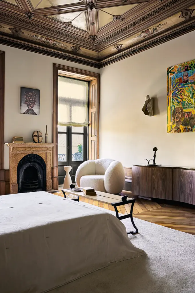 eclectic interior design bedroom with cream walls and with elaborated wooden ceiling and with a marble fireplace and with a wood sideboard and with a bouclé armchair and with a cream rug and with a large bed