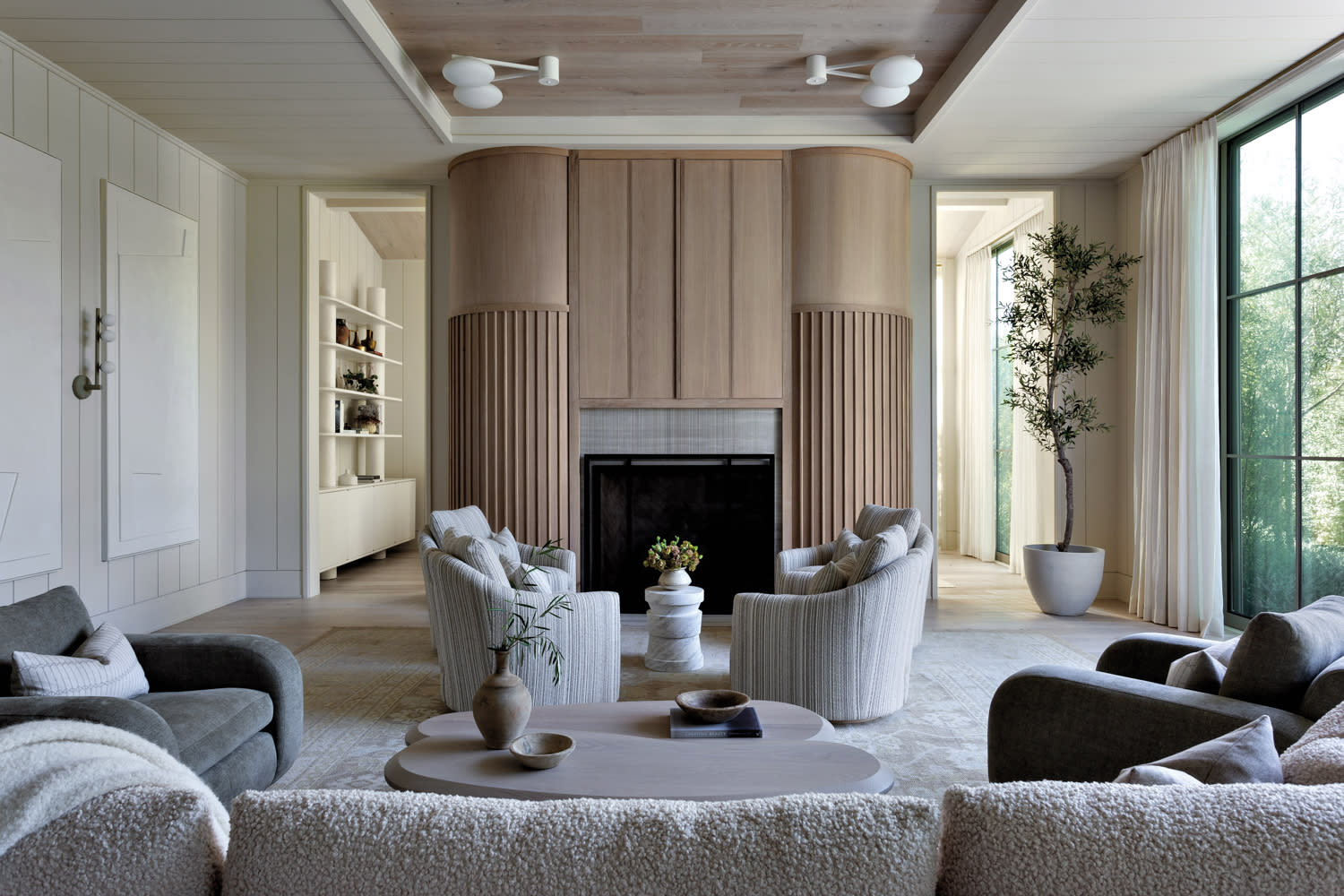 A serene living room with custom creations like a fluted fireplace and unique lighting. Neutral tones, plush seating, and a potted plant add warmth.
