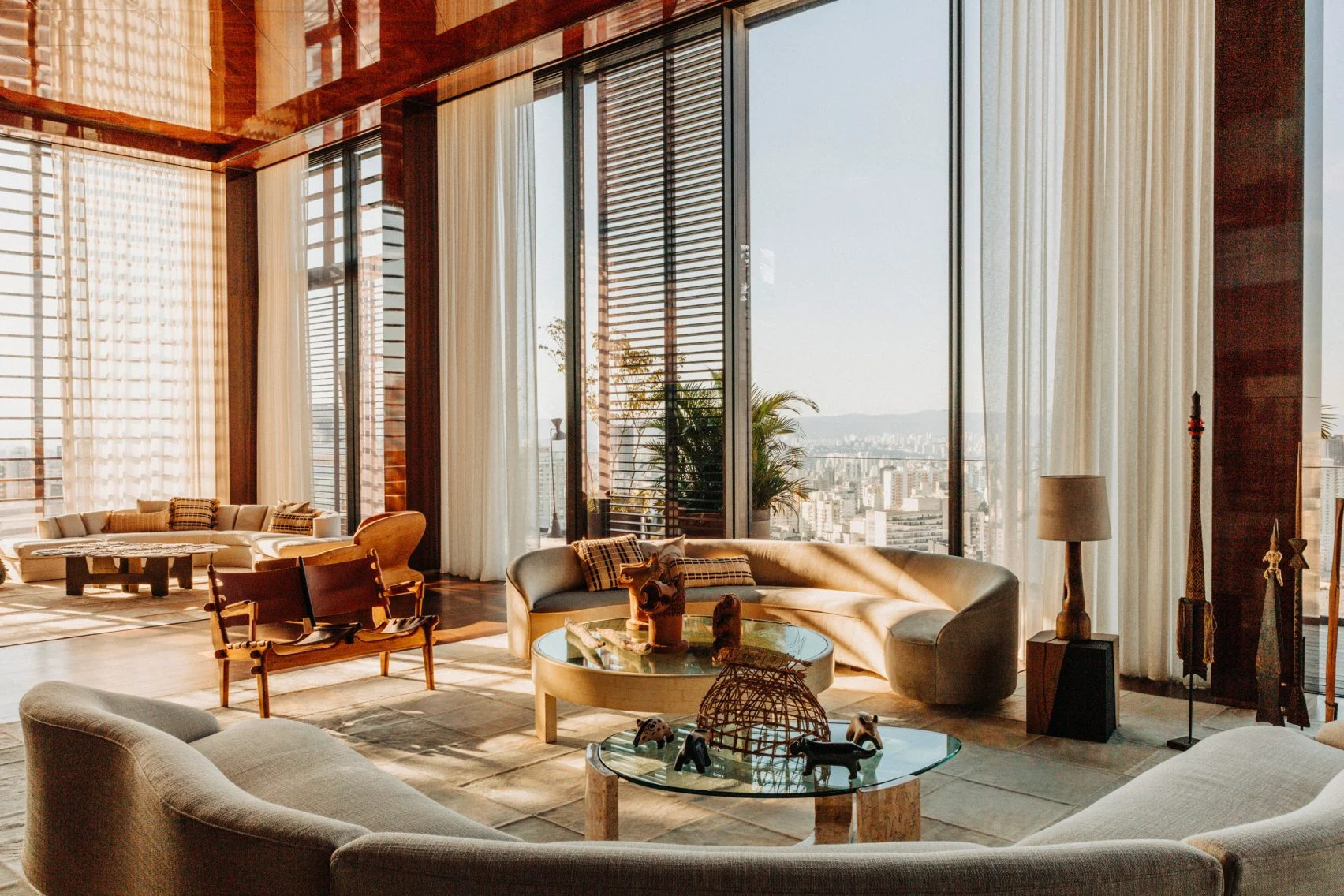 Airy Brazilian Modernism living room with curved sofas, warm wood, and stunning city views through vast windows.