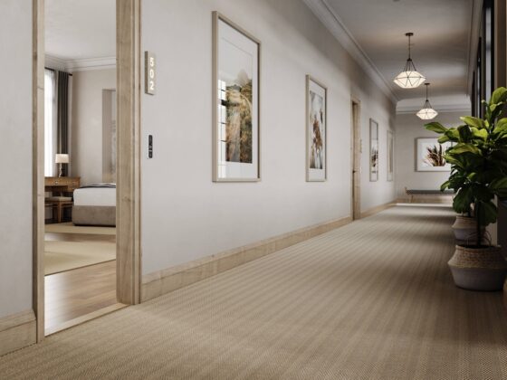 A long hotel hallway with a subtle patterned area rug, framed art, and pendant lights. A glimpse of a room on the left adds depth.