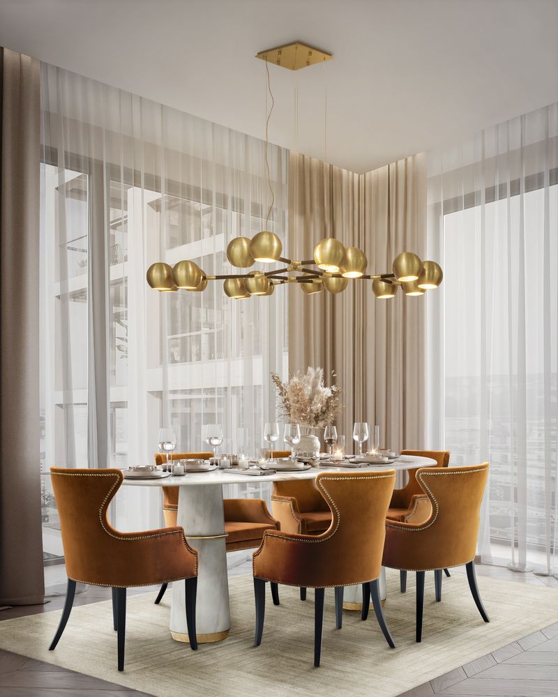 A plush area rug grounds a chic dining room with velvet chairs, a marble table, and a modern chandelier. Sheer curtains soften the bright space.
