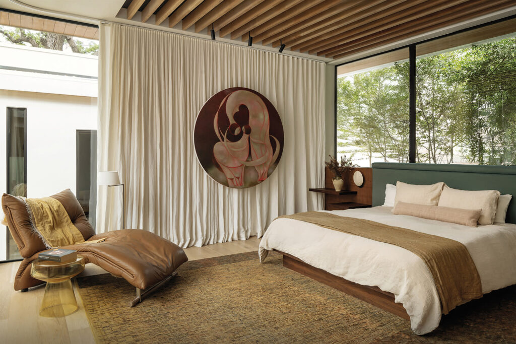 A serene natural home bedroom with a large window overlooking trees, a comfortable chair, and a unique artwork. The bedroom features a mix of textures and natural elements.