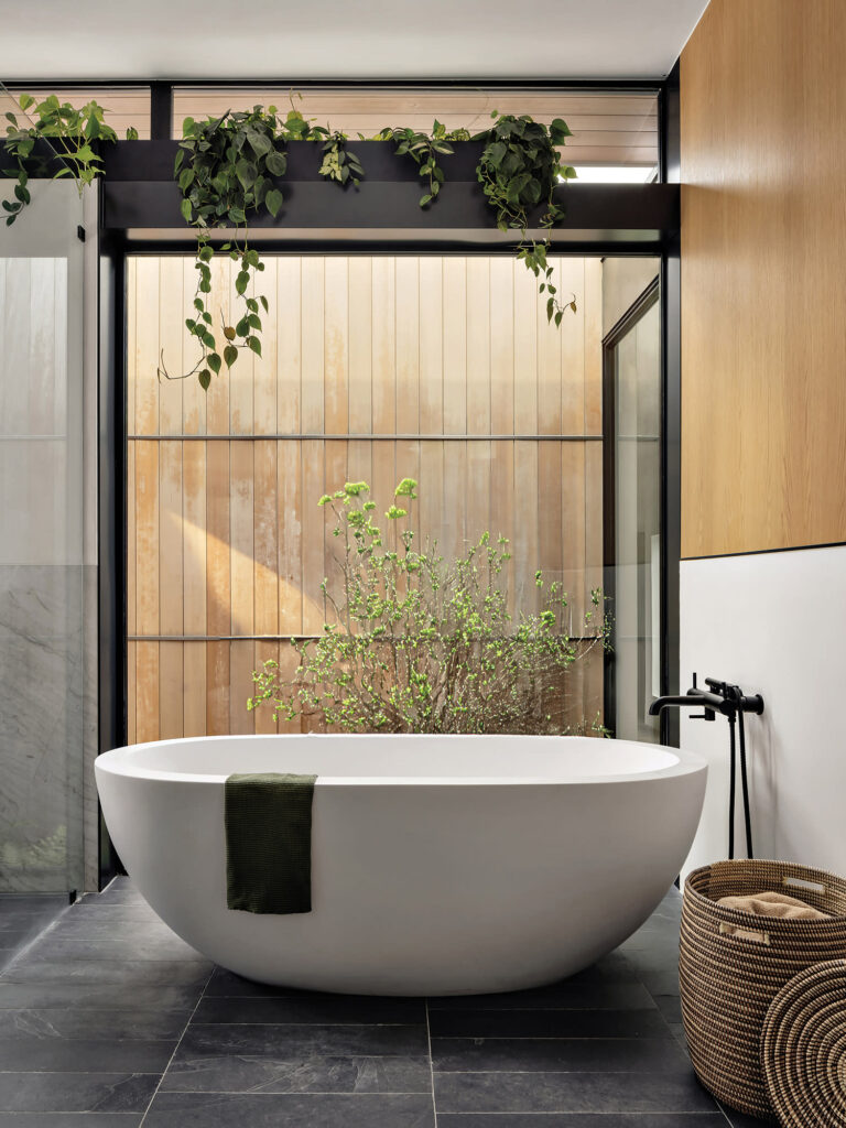 A serene natural home bathroom with a freestanding tub, plants, and a garden view. The bathroom features a mix of natural materials and textures.