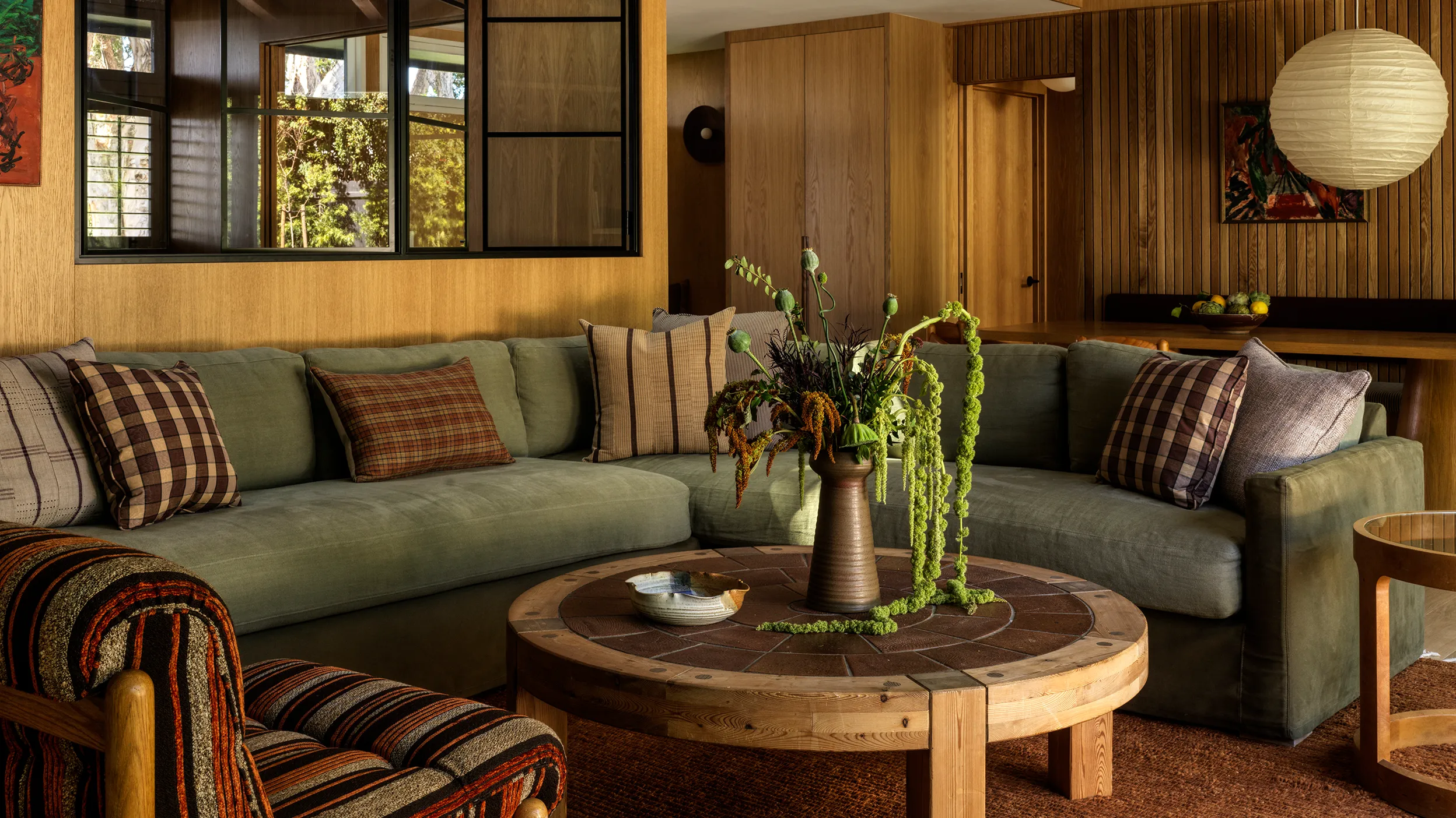 Cozy living room in a wood house with green sofa, round wood table, and striped armchair. Natural light filters through the window.