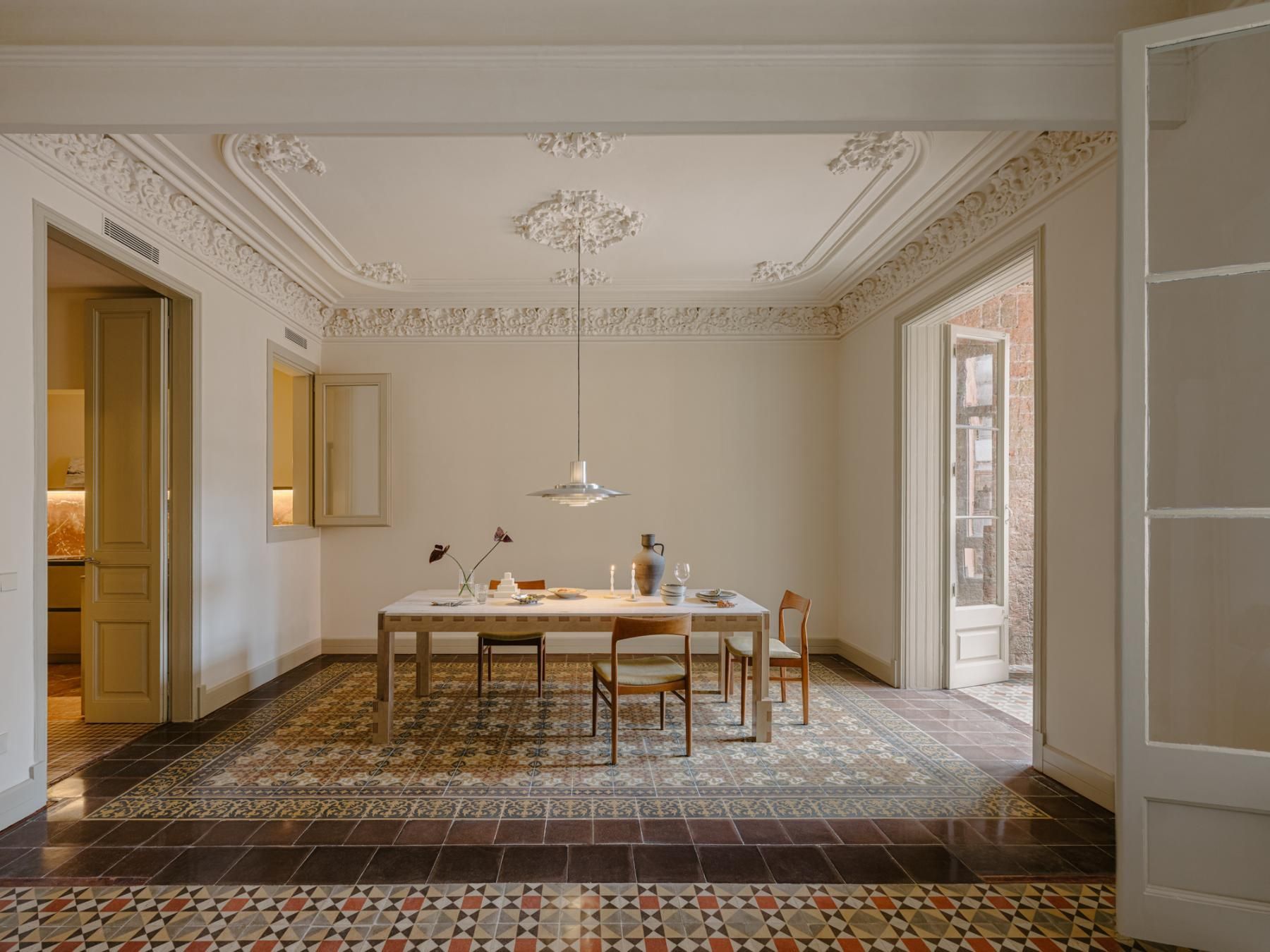 A modern apartment boasts ornate ceiling details juxtaposed with a minimalist dining set. Patterned tile floors add a touch of vintage charm to the bright space.